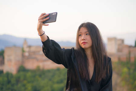 lifestyle portrait of young beautiful and happy Asian Japanese tourist woman having Summer holidays in Europe taking selfie with mobile phone at beautiful Alhambra of Granada in Spainの写真素材