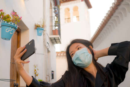 new normal travel and tourism - young happy and beautiful Asian Chinese woman in anti virus mask taking selfie with mobile phone enjoying Summer holidays in Spain after lockdownの写真素材