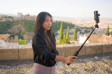 lifestyle portrait of young beautiful and happy Asian Korean tourist woman having Summer holidays in Europe taking selfie with mobile phone at beautiful Alhambra of Granada in Spainの写真素材