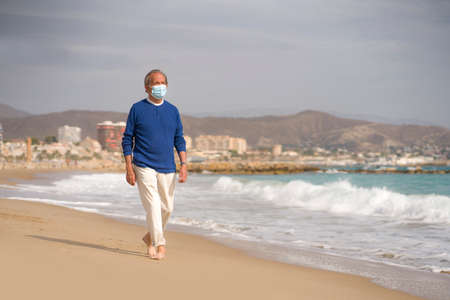 Senior pensioner relaxed in beach during new normal - retired old man on his 70s wearing surgical mask against covid19 walking at the sea thoughtful and contemplative in health conceptの写真素材