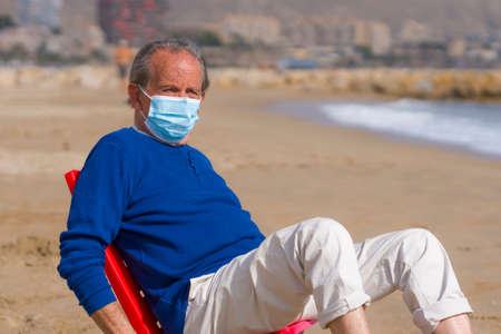 Senior pensioner taking a walk relaxed on the beach - retired old man on his 70s looking at the sea thoughtful and contemplative with surgical mask on his hand in health conceptの写真素材