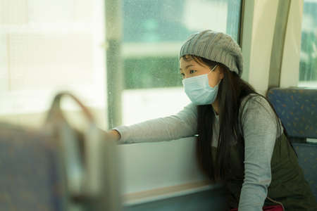 lifestyle portrait of young attractive and pretty Asian woman wearing mask in railcar traveling in new normal virus time - sweet Chinese girl by train window thoughtful and pensiveの写真素材