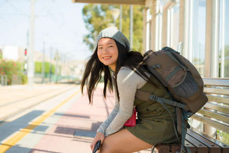 outdoors portrait of young happy and beautiful Asian Korean tourist woman with backpack sitting cheerful on train station bench waiting excited in holidays travel and vacationの写真素材
