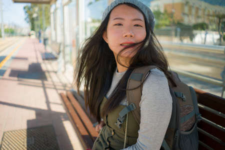 outdoors portrait of young happy and beautiful Asian Japanese tourist woman with backpack sitting cheerful on train station bench waiting excited in holidays travel and vacationの写真素材