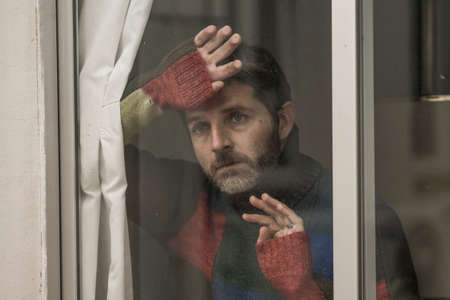 dramatic portrait of attractive sad and depressed mid adult man looking through home window feeling...