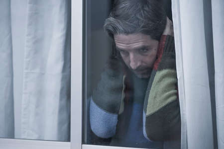 dramatic portrait of attractive sad and depressed mid adult man looking through home window feeling...