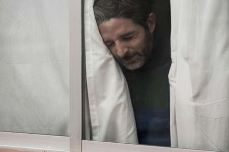 dramatic portrait of attractive sad and depressed mid adult man looking through home window feeling...