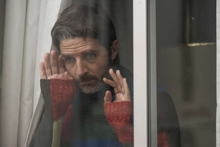 dramatic portrait of attractive sad and depressed mid adult man looking through home window feeling...