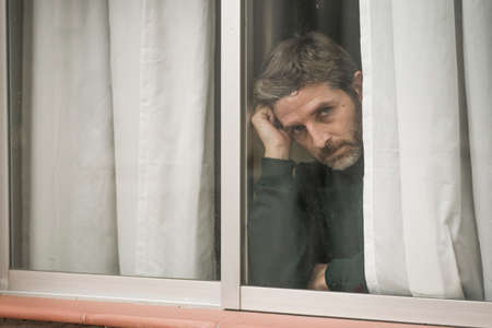 dramatic portrait of attractive sad and depressed mid adult man looking through home window feeling...