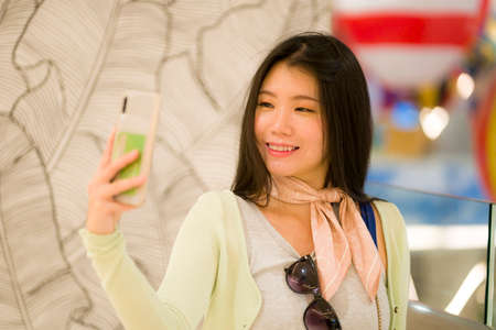 lifestyle portrait of young beautiful and happy Asian Korean woman taking selfie picture with mobile...