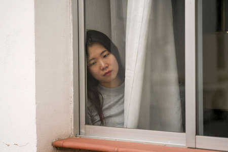 girl in stress at home - dramatic portrait of young sad and depressed Asian Japanese woman on window...