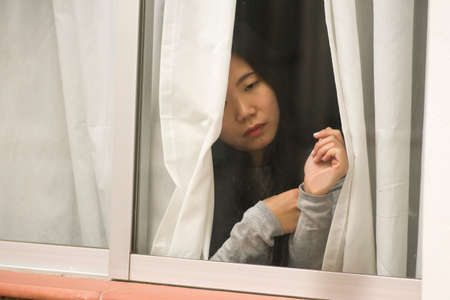 girl in stress at home - dramatic portrait of young sad and depressed Asian Japanese woman on window...