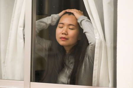 girl in stress at home - dramatic portrait of young sad and depressed Asian Korean woman on window...