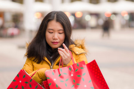 Asian girl enjoying Christmas shopping - young happy and beautiful Japanese woman holding red shopping bag buying presents at street xmas market smiling cheerfulの写真素材