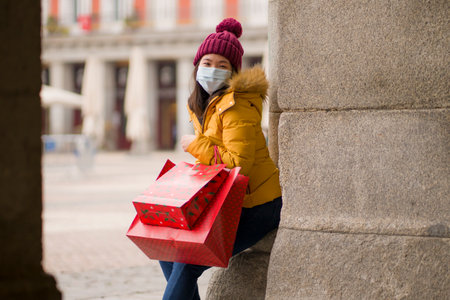 Asian girl enjoying Christmas shopping during  - young happy and beautiful Chinese woman with mask holding red shopping bag buying presents on xmas street marketの写真素材