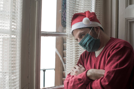 sad and lonely Christmas lockdown during  - depressed and tired man in Santa Claus hat and face mask home alone suffering  restrictions missing familyの写真素材