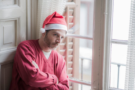 dramatic lifestyle portrait of sad and depressed man in Santa Claus hat feeling lonely home alone by Christmas holiday thoughtful at the window in unhappy xmas conceptの写真素材