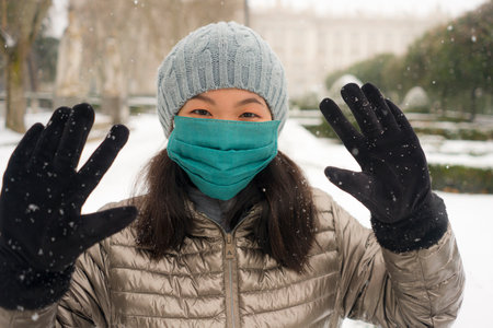 snowfall in covid19 time - young happy and attractive Asian Korean woman in winter hat and mask at beautiful city park playing with snow cheerful enjoying cold Christmas holidaysの写真素材