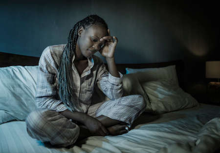 dramatic home portrait of young sick and depressed black afro American girl sitting on bed  upset and sleepless at night feeling overwhelmed suffering depression and anxiety problemの写真素材