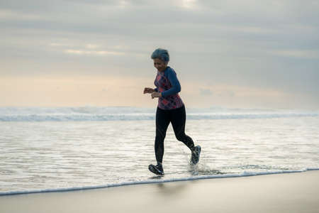 fit and attractive mature woman with grey hair doing beach workout on her 50s running on the beach...