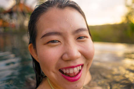 young woman relaxed at luxury resort infinity pool - happy and cheerful Asian Korean girl taking selfie photo in bikini enjoying jungle view at hotel swimming pool during summer holidays travelの写真素材