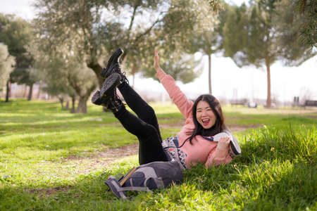 lifestyle portrait of young happy and beautiful Asian Korean woman in pink hoodie lying on green grass cheerful and excited playing outdoors enjoying carefreeの写真素材