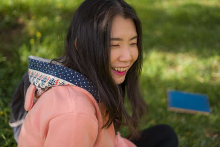 sweet Asian woman smiling relaxed outdoors - lifestyle portrait of young happy and beautiful Japanese girl sitting playful on green grass in city park smiling cheerfulの写真素材