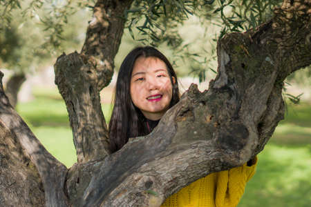 pretty Asian woman in city park - lifestyle portrait of young beautiful and happy Chinese girl playful by a tree enjoying enjoying outdoors cheerful and relaxedの写真素材