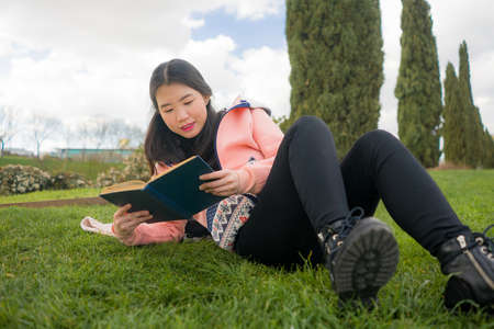 young Asian woman enjoying novel on grass - lifestyle portrait of young happy and pretty Japanese girl reading a book at beautiful city park in reading and studying conceptの写真素材