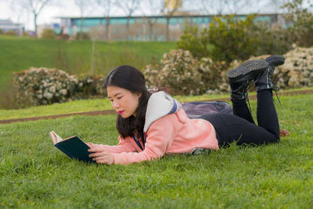 young Asian woman enjoying novel on grass - lifestyle portrait of young happy and pretty Korean girl reading a book at beautiful city park in reading and studying conceptの写真素材