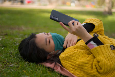 young Asian woman in city park  - outdoors lifestyle portrait of happy and pretty Chinese girl in face mask using internet with mobile phone relaxed on green grassの写真素材