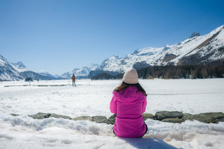 woman enjoying amazing snowy landscape view - back of girl in winter jacket sitting in front of frozen lake and snow mountains enjoying Swiss Alps getawayの写真素材