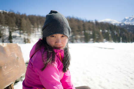 winter outdoors lifestyle portrait of young happy and attractive Asian Korean woman enjoying on bench in snow at beautiful alpine mountain during Christmas holidayの写真素材