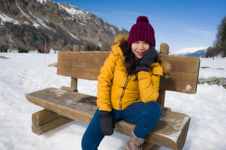 young happy and beautiful Asian Chinese woman in yellow jacket and winter hat smiling cheerful on bech over frozen lake covered in snow in Swiss Alps mountainsの写真素材