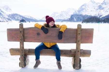 young happy and beautiful Asian Chinese woman in yellow jacket and winter hat smiling cheerful on bench over frozen lake covered in snow in Swiss Alps mountainsの写真素材