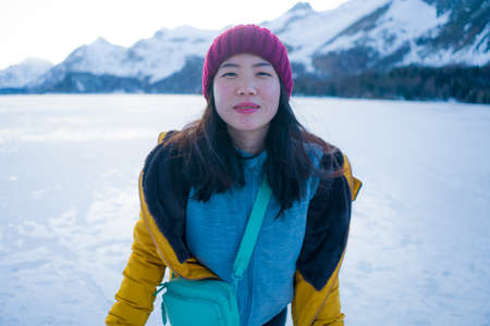 lifestyle portrait of young happy and beautiful Asian Korean woman in winter jacket and beanie enjoying the snow standing on frozen lake in Swiss Alps mountainの写真素材