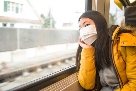 lifestyle portrait of young beautiful and thoughtful Asian Korean woman in face mask as protection vs looking through window during train tripの写真素材