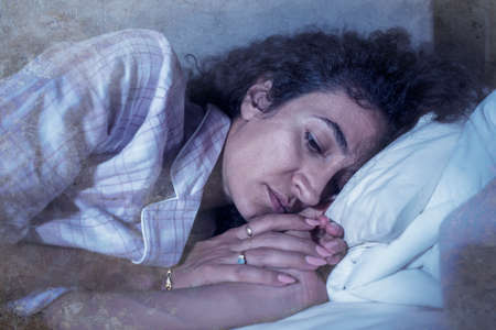 dark and edgy portrait of depressed and sleepless latin woman lying worried and awake on bed at night suffering insomnia and depression problemの写真素材