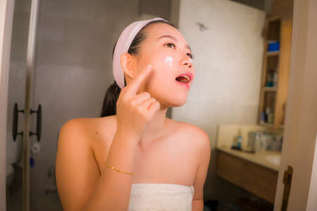 portrait of young happy and beautiful Asian Chinese woman applying facial anti wrinkle cream and serum at bathroom mirror in skin care and beauty treatment conceptの写真素材