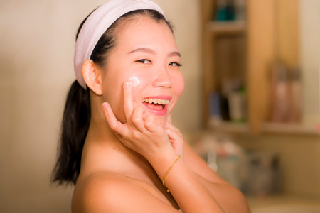 portrait of young happy and beautiful Asian Japanese woman applying facial anti wrinkle cream and serum at bathroom mirror in skin care and beauty treatment conceptの写真素材