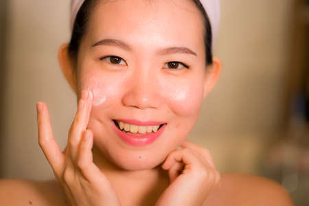portrait of young happy and beautiful Asian Chinese woman applying facial anti wrinkle cream and serum at bathroom mirror in skin care and beauty treatment conceptの写真素材