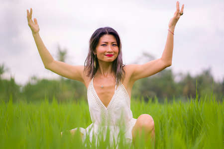 artistic portrait of young attractive and happy Asian woman outdoors at green rice field landscape wearing elegant long dress enjoying playful on beautiful natureの写真素材