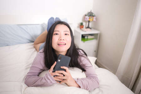 Asian girl with hand phone - home lifestyle portrait of young happy and attractive Chinese woman on...