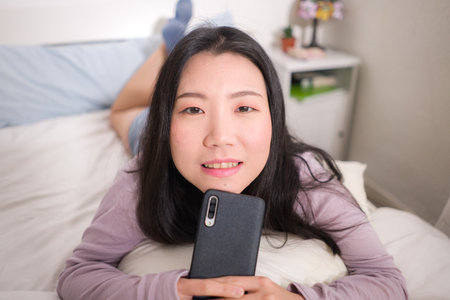 Asian girl with hand phone - home lifestyle portrait of young happy and attractive Japanese woman on bed using internet mobile internet app smiling cheerful in bedroomの写真素材