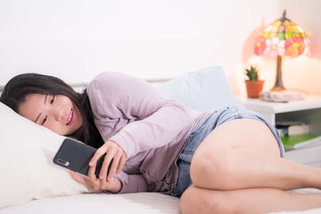 Asian girl with hand phone - home lifestyle portrait of young happy and attractive Korean woman on bed using internet mobile internet app smiling cheerful in bedroomの写真素材