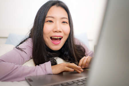 home lifestyle portrait of young Asian woman networking on bed -  beautiful and happy Korean girl in bedroom using laptop computer smiling relaxed and cheerfulの写真素材