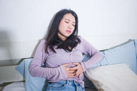 young Asian woman suffering menses abdominal cramp - sick and tired Chinese girl lying on bed having having period pain holding her stomach feeling unwellの写真素材