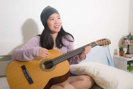 young happy Asian woman playing guitar in bed - attractive and beautiful Korean girl in bedroom enjoying singing and writing music relaxed at homeの写真素材