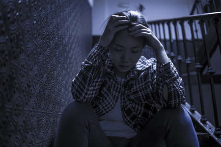 young beautiful Asian woman in pain suffering depression - dramatic indoors portrait on staircase of sad and depressed Chinese girl as victim of bullying and abuseの写真素材