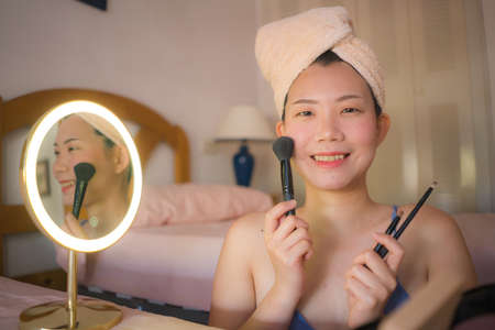 pretty Asian woman applying facial makeup - lifestyle portrait of young happy and beautiful Chinese girl at home brushing her face applying beauty treatment in mirrorの写真素材